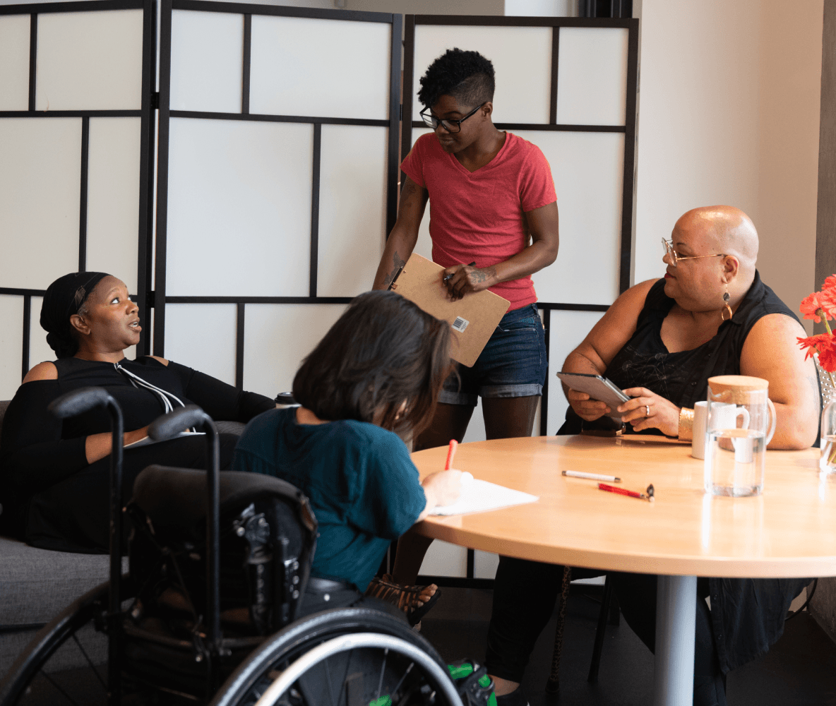 Four disabled people of color gather around a table during a meeting. A Black woman sitting on a couch speaks with a neutral expression while the three others (a South Asian person sitting in a wheelchair and taking notes, a Black non-binary person sitting in a chair with a tablet and cane, and a Black non-binary person standing with a clipboard) listen.